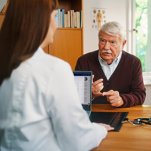 Ärztin mit Laptop im Gespräch mit einem älteren Patienten.