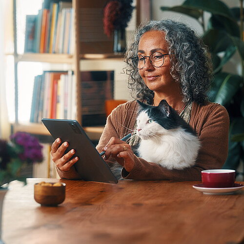 Frau sitzt mit Katze auf dem Schoß am Wohnzimmertisch und tippt mit Stick auf Tablet.