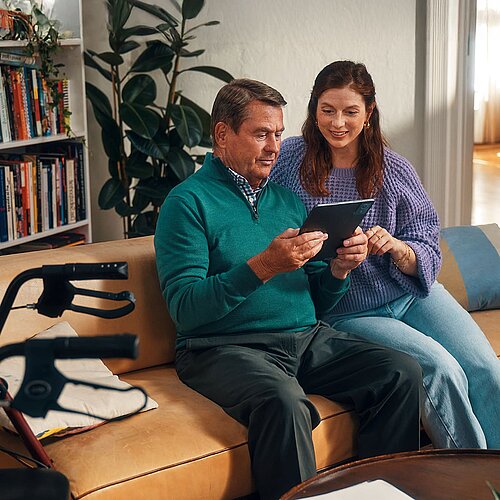 Älterer Mann und jüngere Frau sitzen auf dem Sofa und schauen zusammen auf ein Tablet. 