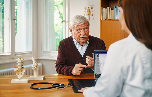 Ärztin mit Laptop im Gespräch mit einem älteren Patienten.