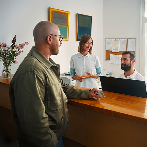 Patient mit Versichertenkarte spricht mit Mitarbeitenden am Empfang einer Arztpraxis. 